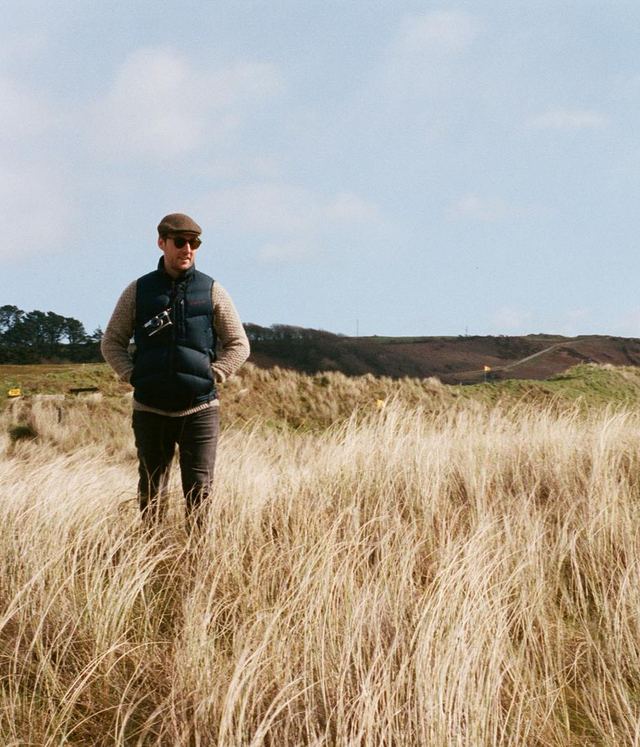 Ray O'Neill standing in tall dune grass under a bright sky, wearing a flat cap, gilet and camera