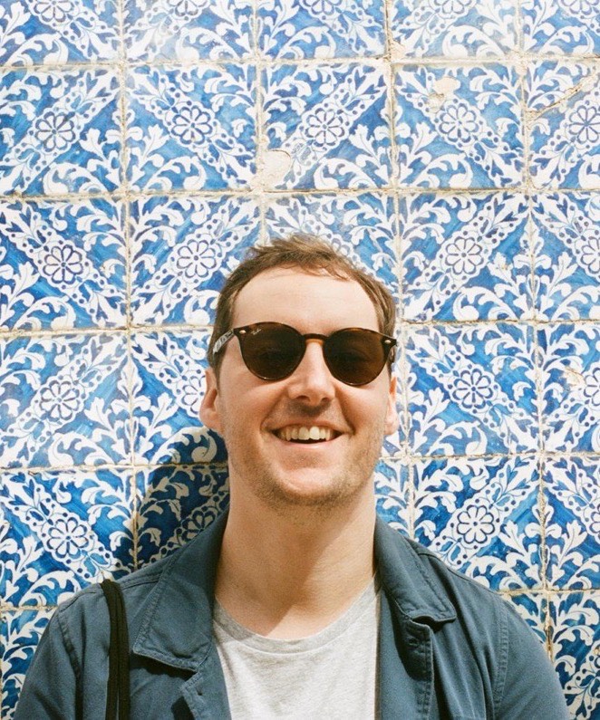 Ray O'Neill, smiling in sunglasses and a denim jacket, in front of a wall of blue and white patterned tiles